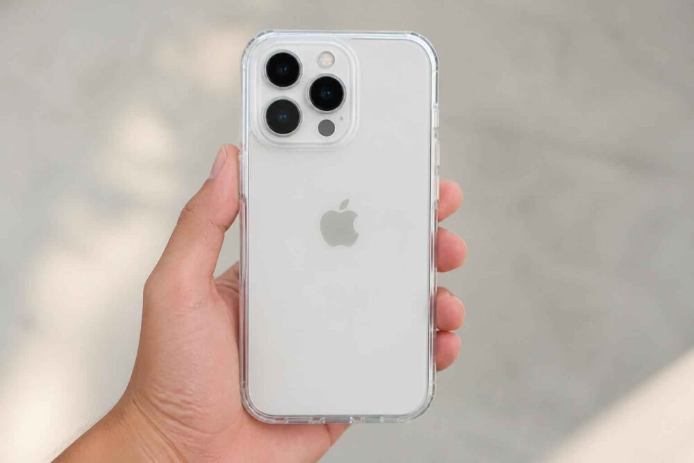 Biały iPhone trzymany w dłoni Biały iPhone z potrójnym aparatem w przezroczystym etui trzymany na tle jasnej powierzchni